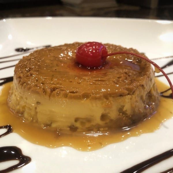 A flan dessert with caramel sauce, chocolate drizzle, and a maraschino cherry on a white plate.