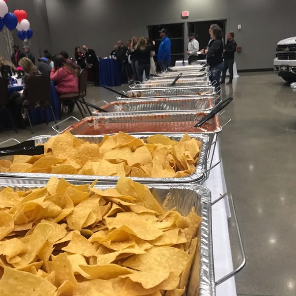 A buffet line with large trays of tortilla chips and salsa, with people in the background.