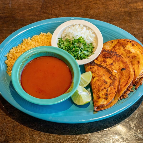 Los Mariachis birria tacos with consommé, rice, diced onions, cilantro, and lime wedges on a blue plate.