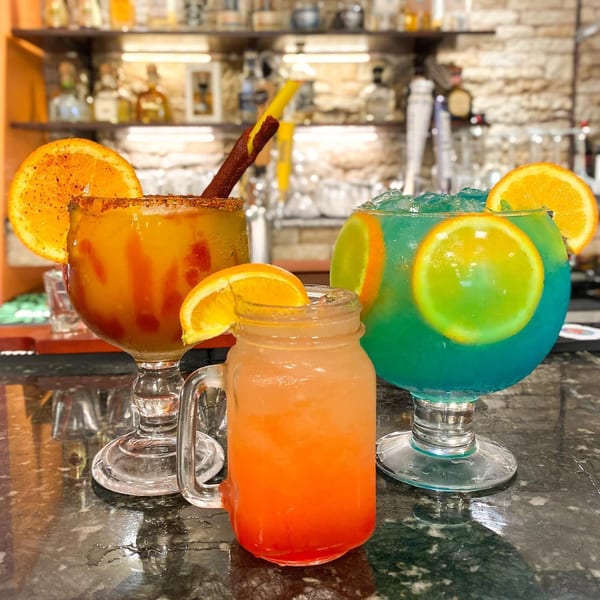 Three colorful Mexican cocktails, two margaritas and one mason jar drink, garnished with orange slices on a bar.