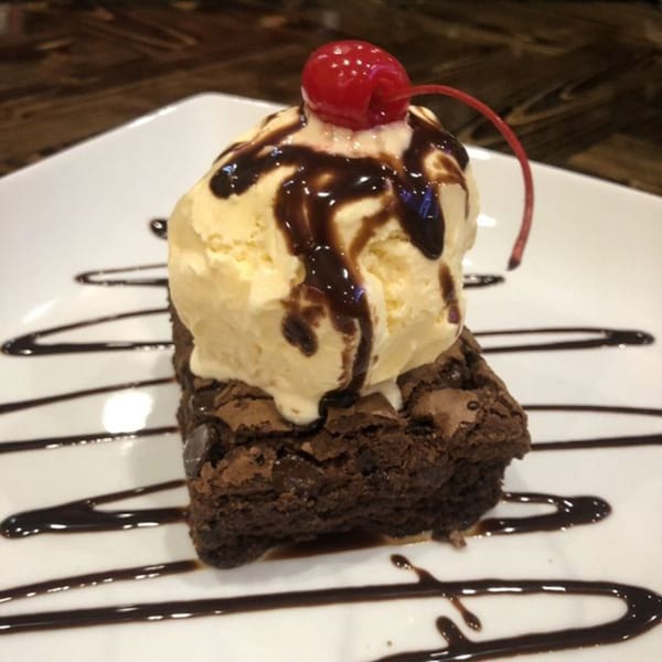 A brownie topped with vanilla ice cream, chocolate syrup, and a maraschino cherry on a white plate.