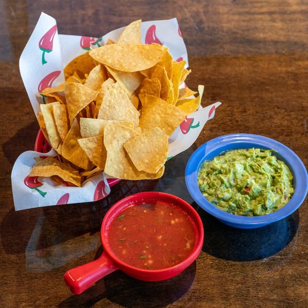 A basket of tortilla chips with a side of guacamole and a bowl of salsa on a wooden table.