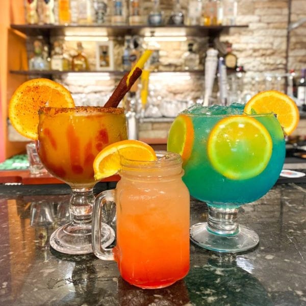 Three colorful Mexican drinks, two margaritas and one layered cocktail, on a bar counter.