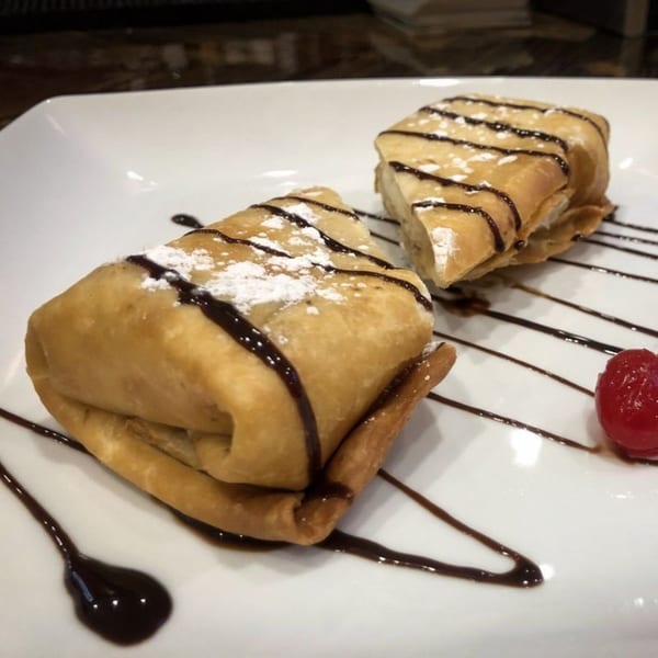 Two golden-fried chimichangas drizzled with chocolate syrup and powdered sugar, with a cherry on a white plate.